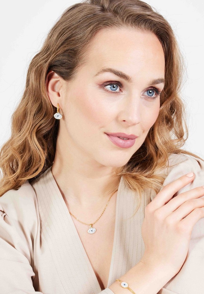 Goudkleurige sieradenset met een ovale hangerketting, bijpassende oorbellen en een armband met blauwe accenten, gepresenteerd op een model.