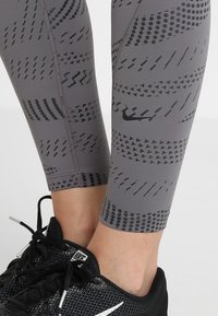 Leggings atletici grigi presentano un motivo geometrico con punti e linee, realizzati in materiale elastico, abbinati a scarpe sportive nere.