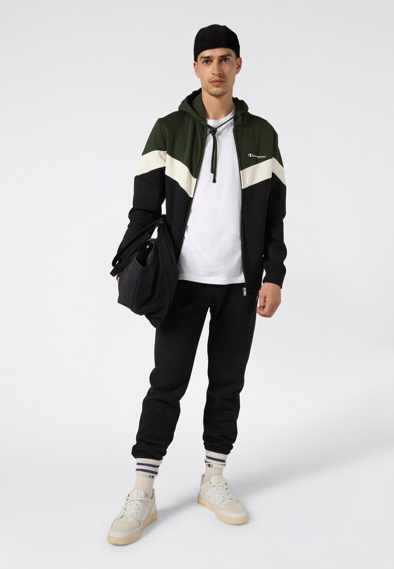 Uomo in felpa nera e verde con zip, t-shirt bianca, pantaloni neri, sneaker bianche e cappellino nero in piedi, che tiene un sacchetto nero in una mano.