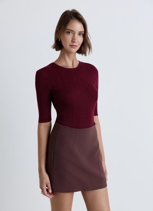 Donna con capelli castani corti che indossa un top bordeaux a coste con maniche al gomito e una gonna marrone scuro a vita alta su uno sfondo semplice.