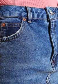 Džínová sukně v středně modré barvě, s třepeným lemem, standardními kapsami a kontrastním prošitím; těsně přiléhající pas a klasický pětikapsový design.