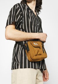 Persona che indossa una camicia a maniche corte a righe bianche e nere, pantaloni beige e una piccola borsa a tracolla marrone con tasche frontali.