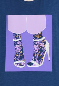 Grafické tričko s designem fialově vzorovaných ponožek v kombinaci se stříbrnými vysokými podpatky na fialovém pozadí.