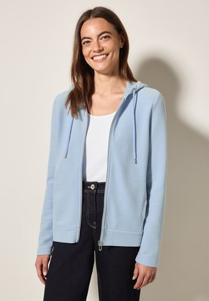 Haut de sweat à capuche zippé bleu clair en tissu texturé, avec un capuchon à cordon de serrage et des poignets côtelés. Associé à un haut blanc et un jean foncé.
