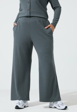 Osoba ubrana w szare prążkowane szerokie spodnie i dopasowany zapinany top z rękami w kieszeniach, zestawiona z białymi sneakersami New Balance.