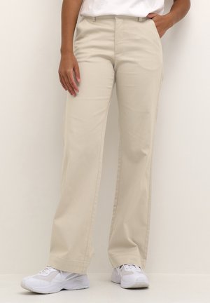 Osoba ubrana w beżowe spodnie o prostych nogawkach, białe buty sportowe i biały top, stojąca na tle jasnego, jednolitego tła.