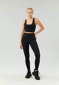 Svart sportbh og høytlivede leggings laget av elastisk, glatt stoff. Modell iført svarte treningssko. Ensfarget grå bakgrunn.
