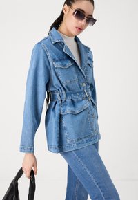 Lichtblauwe spijkerjas met een ceintuur in de taille, knoopsluiting en vier voorzakken, gecombineerd met bijpassende spijkerbroek en zonnebril.