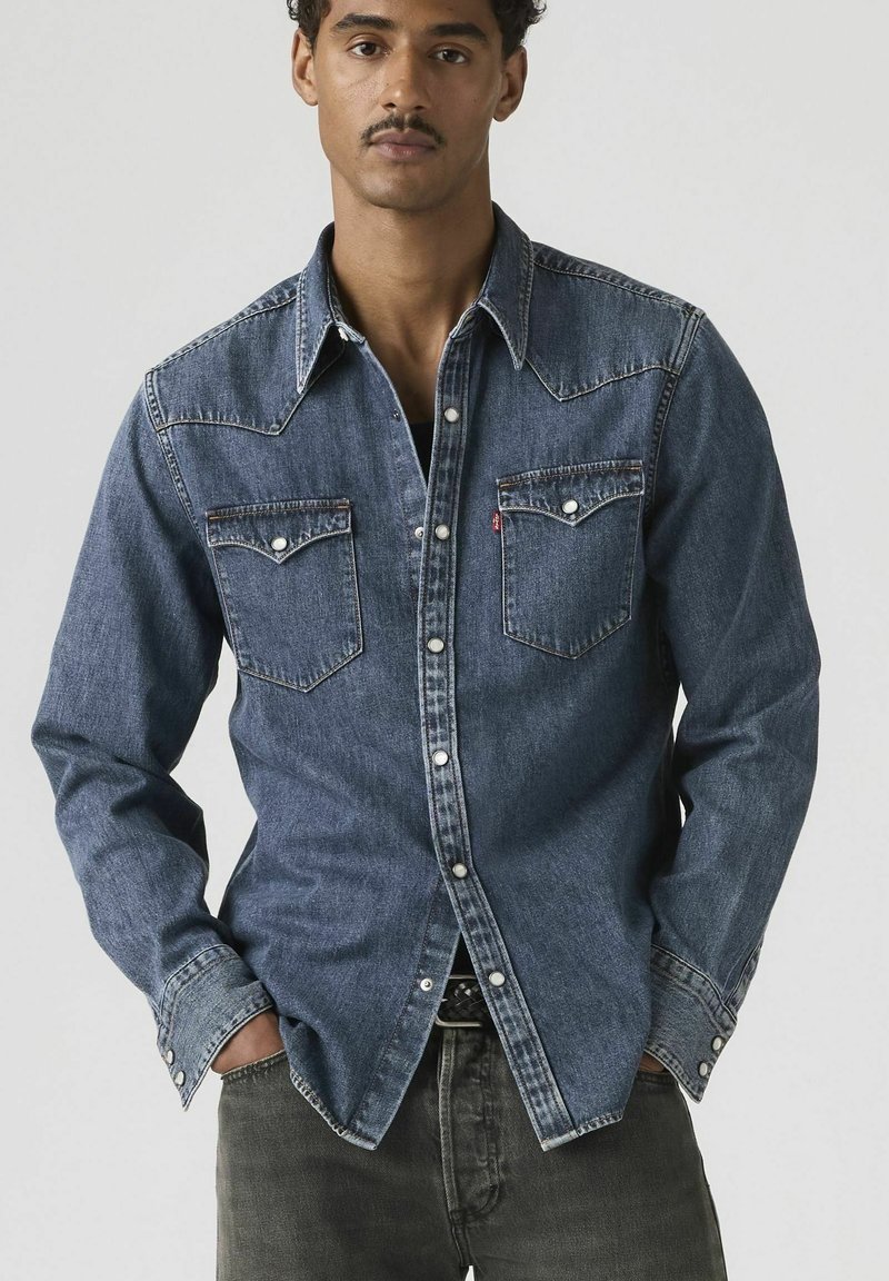 Mann trägt ein geknöpftes blaues Jeanshemd mit Brusttaschen, steht mit den Händen in den Taschen der dunklen Hose vor schlichtem Hintergrund.