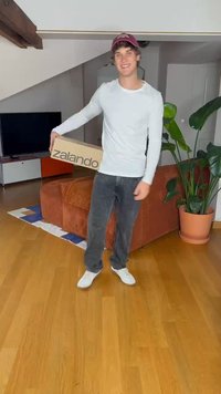 Zalando cardboard box held by a person in a light long-sleeve shirt and dark jeans, standing on wooden flooring with a plant nearby.
