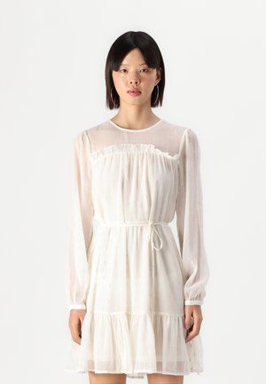 Robe blanche avec des manches longues transparentes, un col froncé, une ceinture à la taille et un ourlet en étages. Fabriquée dans un tissu texturé avec de petites accents réflecteurs.