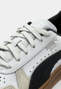 Sneaker bianco in pelle con design perforato, dettagli in suede nero e suola in gomma beige; include lacci piatti e dettaglio del logo.