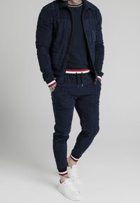 Marineblauwe ritssluiting jack en joggers met reliëftekst ontwerp. Witte en rode gestreepte manchetten. Gestyled met witte sneakers voor een casual look.
