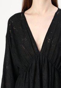 Μαύρη δαντελένια μπλούζα με V λαιμόκοψη, πλούσιες λεπτομέρειες στη μέση και μακριά μανίκια με υφασμάτινη, ημιδιάφανη υφή.