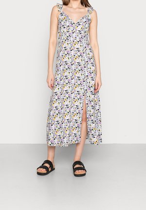 Žena v květovaných midi šatech bez rukávů s volánkovými ramínky a předním rozparkem, doplněná černými sandály.