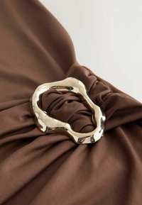 Bruine stof met een gedraaid ontwerp, voorzien van een prominente metallic zilveren lus als accent. De textuur is glad, wat de val benadrukt.