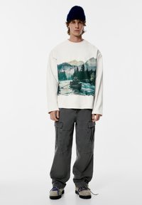 Bílá mikina s potiskem krajiny, doplněná šedými kalhotami cargo. Model nosí modrou čepici a texturované šedé boty.