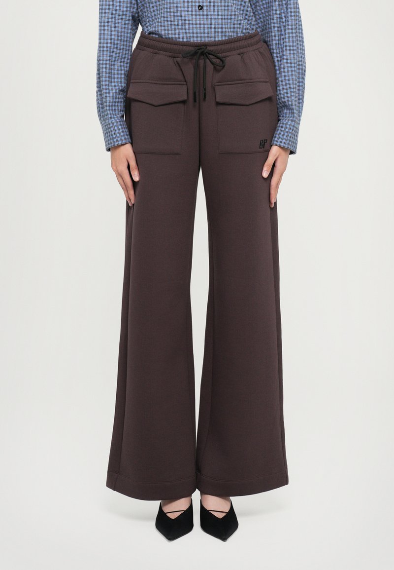 Καφέ παντελόνι σε φαρδιά γραμμή με μεγάλες μπροσ�τινές τσέπες με καπάκι, ελαστική μέση με κορδόνι, φορεμένο με μαύρες γόβες και μπλε καρό πουκάμισο.