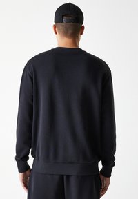 Sudadera negra de tela suave, con cuello redondo, puños y dobladillo acanalados. Diseño liso, sin logotipos ni patrones visibles.