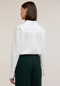 Camicia bianca a maniche lunghe con colletto, dettaglio plissettato sul retro e polsini con bottoni, abbinata a pantaloni su misura verde scuro.