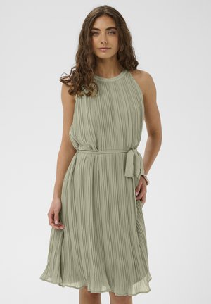 Femme aux cheveux bruns bouclés portant une robe midi sans manches plissée vert clair avec une ceinture nouée à la taille, debout devant un fond uni.