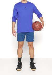 Långärmad klarblå skjorta med svart logotyp, parat med blå mesh shorts med vita Accenter, och svarta sportskor. Håller en basketboll.