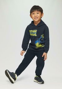 Marineblauwe hoodie met "STAY COOL" grafiek en blauwe accenten, gecombineerd met bijpassende marineblauwe broek. Atletische schoenen hebben een donkere bovenkant en groene accenten.