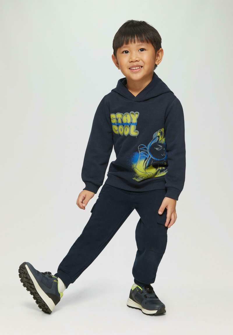 Marineblauwe hoodie met "STAY COOL" grafiek en blauwe accenten, gecombineerd met bijpassende marineblauwe broek. Atletische schoenen hebben een donkere bovenkant en groene accenten.