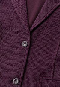 Gros plan d'une veste texturée violet foncé avec deux boutons visibles et une poche avant, montrant les détails du col et du revers.