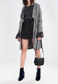 Un manteau long gris sur un pull côtelé noir, une mini-jupe noire à boutons, un sac bandoulière noir et des bottines bleues.