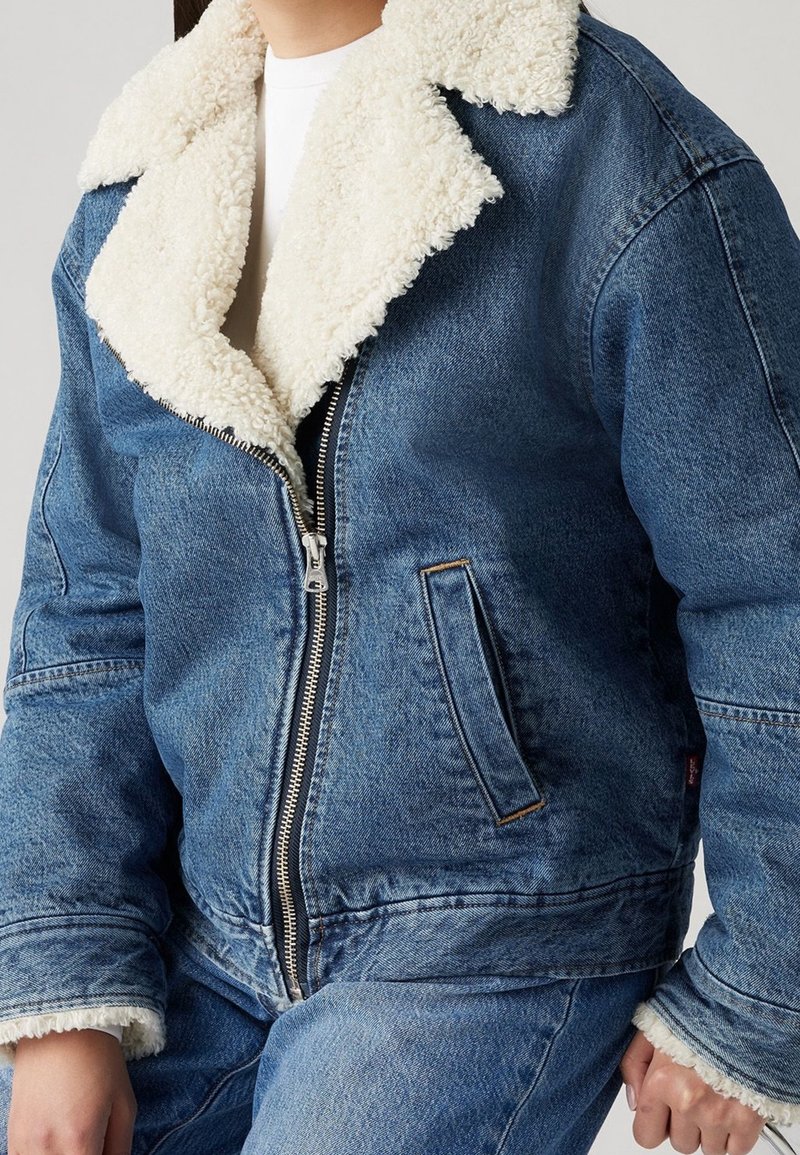 Personne portant une veste en denim bleu avec doublure sherpa blanche, fermeture éclair devant, et pantalon en denim bleu assorti, sur fond uni.