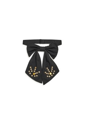 Nœud papillon en tissu noir avec deux longues queues décorées de perles et de clous dorés disposés.