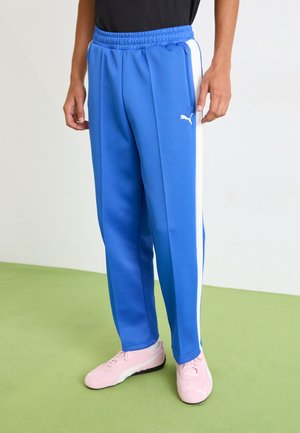 Blue athletic pants with white side stripes, made of a smooth fabric, featuring an elastic waistband and zip pockets. Paired with pink sneakers.
