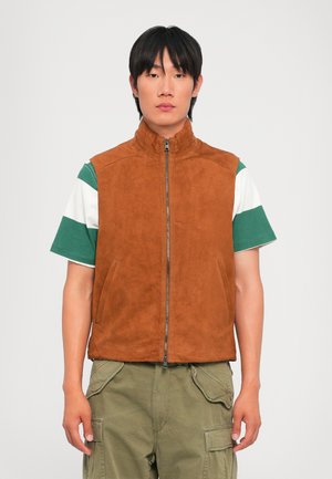 Braune Wildlederweste mit hohem Kragen und Frontreißverschluss, kombiniert mit einem grün-weißen gestreiften T-Shirt und olivfarbener Cargo-Hose.