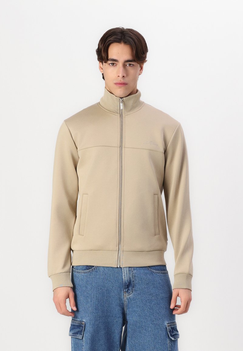 Beige Zip-Jacke mit hohem Kragen, ausgestattet mit zwei Fronttaschen und einer glatten Textur. Getragen mit blauen Denim-Shorts.