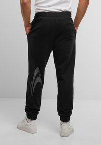 Sorte sweatpants med elastisk talje, sidelommer og et gråt abstrakt mønster på venstre ben. Kanteprofiler ved anklen.