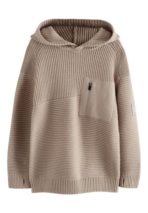 Chlapec s kučeravými vlasmi v béžovej kapucni, čiernych cargo nohaviciach a bielych teniskách stojí pri kamennej stene na mestskom chodníku.