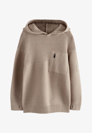 Garçon aux cheveux bouclés portant un sweat à capuche beige, un pantalon cargo noir et des baskets blanches, debout contre un mur en pierre sur un trottoir en ville.