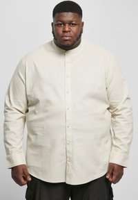 Beige blouse met een Mandarin-collet, lange mouwen en een zachte textuur; heeft een rechte zoom en lichtgekleurde knopen.