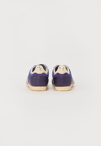 Des chaussures de sport violettes avec une tige texturée, des accents crème et une semelle en caoutchouc. Elles présentent trois rayures sur les côtés et un logo sur le talon.