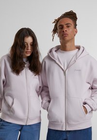 Felpe con cappuccio rosa chiaro, con zip, tasche frontali e orlo aderente. I modelli mostrano una vestibilità comoda, con il testo ricamato "Proibito" sul petto.