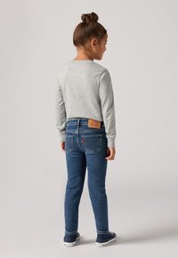 Dijete s punđom nosi sivi duksericu s dugim rukavima, plave traperice i tamnoplave tenisice, okrenuto leđima prema običnoj pozadini.