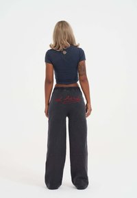 Žena se světle hnědými vlasy, oblečená v tmavém crop topu s designem růže a černých kalhotách s širokými nohavicemi, na zadní straně s červeným textem "ed hardy".