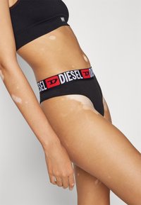 String noir avec une ceinture logo "DIESEL" rouge et blanche, présentant une texture de tissu lisse et un design minimaliste.