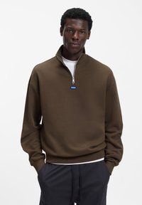 Rjava jopa z zadrgo z visokim ovratnikom in modrim logotipom na prsih, z dolgimi rokavi in sproščenim krojem.