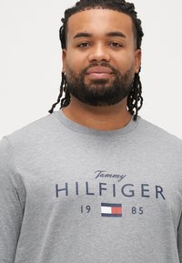 Szara koszulka z długim rękawem z granatowym logo "Tommy HILFIGER" z rokiem "1985" i trójkolorowym emblematem poniżej. Gładka tekstura.