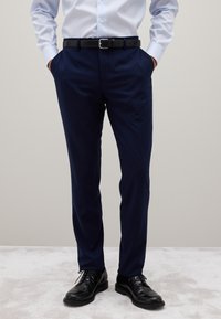 Mann in navyblauen Anzughosen, schwarzem Gürtel, schwarzen Lederschuhen und hellblauem Hemd mit Knöpfen, die die Hände in den Taschen hat.