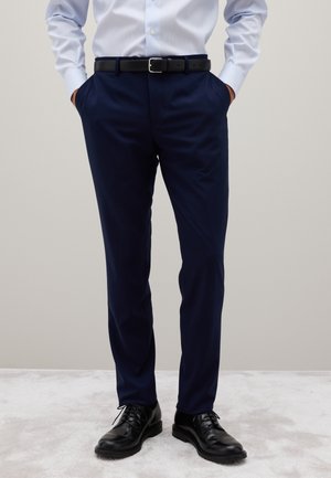 Hombre que lleva pantalones de vestir azul marino, cinturón negro, zapatos de vestir de cuero negro y camisa de botones azul clara con las manos en los bolsillos.