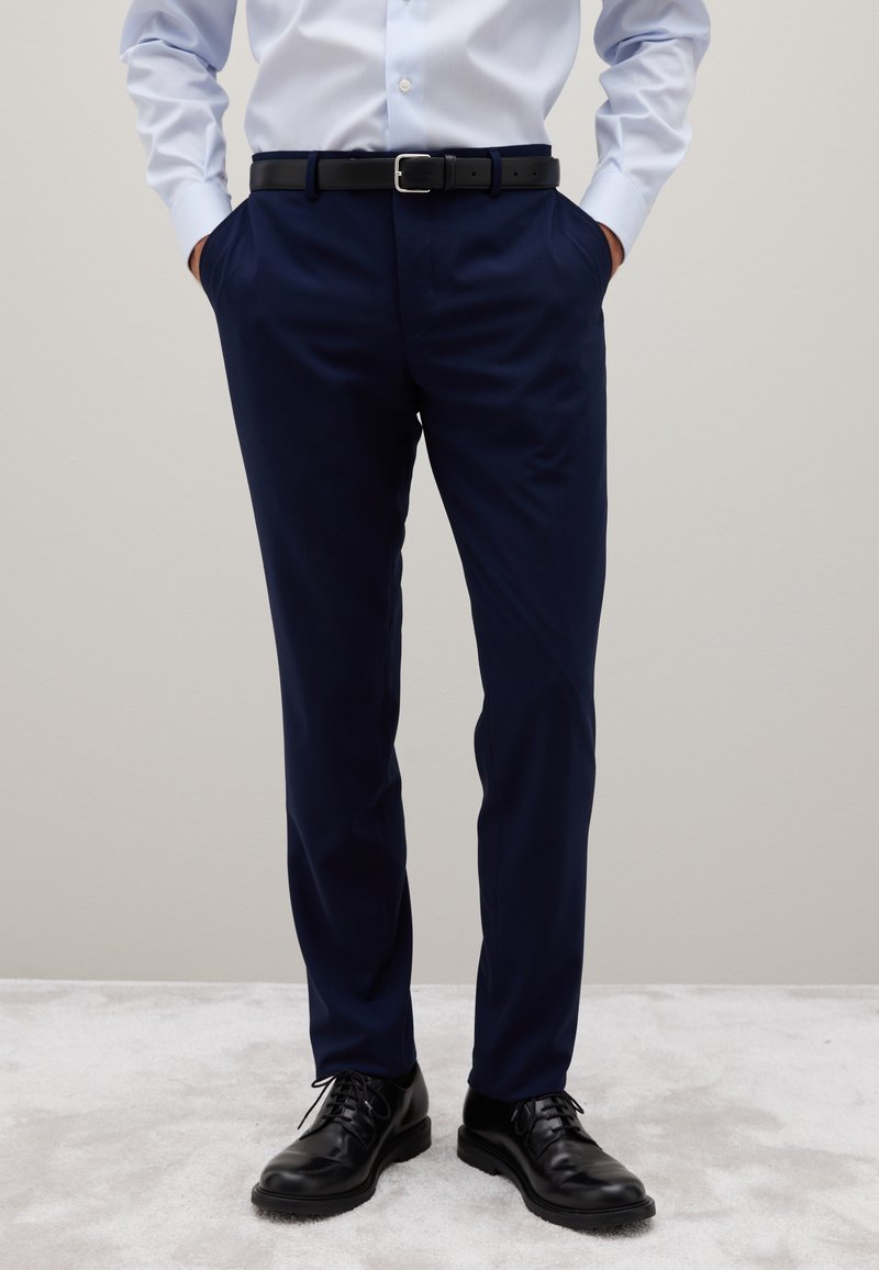 Mann in navyblauen Anzughosen, schwarzem Gürtel, schwarzen Lederschuhen und hellblauem Hemd mit Knöpfen, die die Hände in den Taschen hat.