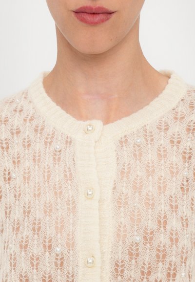 Cardigan di colore crema chiaro con motivo di pizzo, scollo rotondo e chiusura a bottone. Decorato con piccole perle in tutta la sua superficie.